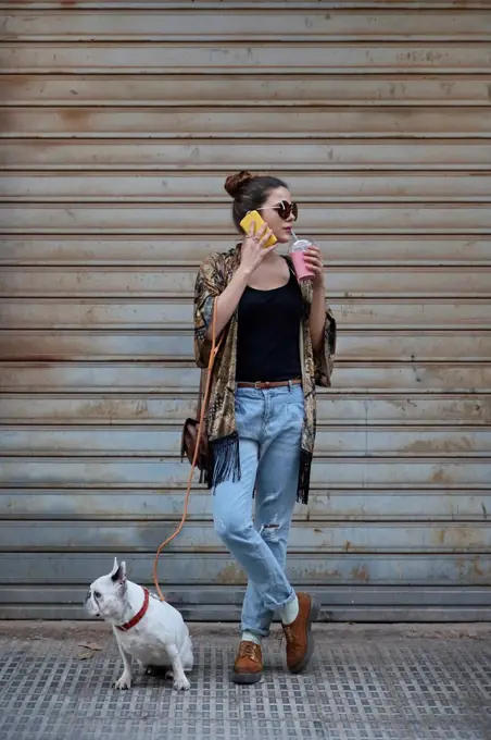 Young woman drinking smoothie and telephoning with smartphone while waiting with her dog on the street