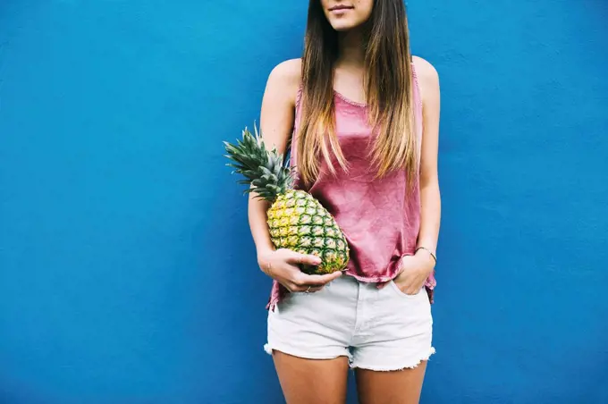 Young woman standing with pineapple at blue wall