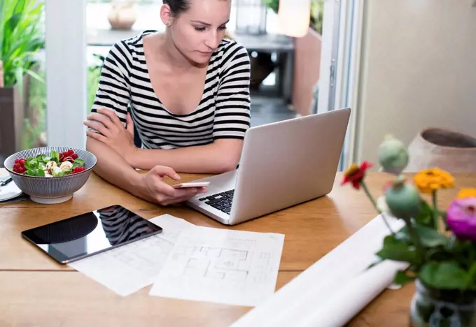 Woman at desk using laptop and cell phone next to construction plan and salad