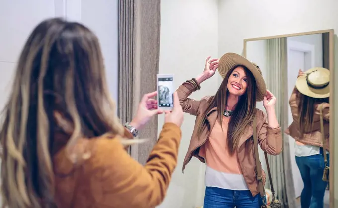 Woman taking picture of friend putting on straw hat in a boutique