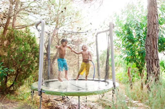 Two little boys jumping on trampoline while splashing with water from garden hose