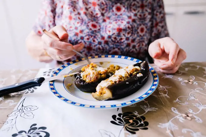 Senior woman eating stuffed eggplant in the kitchen, partial view