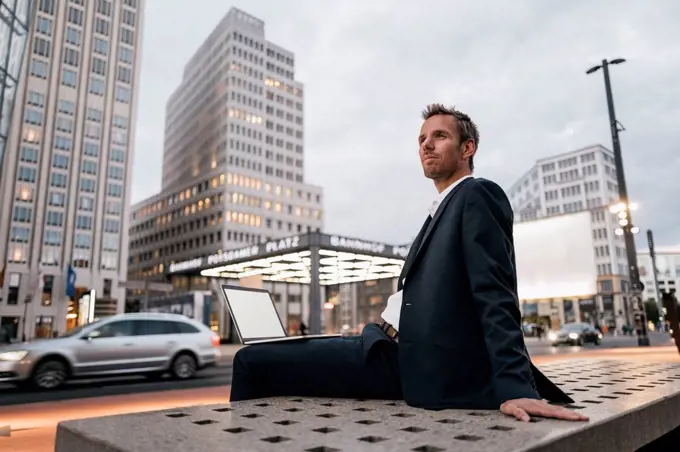 Germany, Berlin, Potsdamer Platz, businessman sitting on bench with laptop in the evening