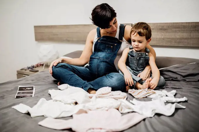 Pregnant woman and her little son sitting together on bed with baby clothing