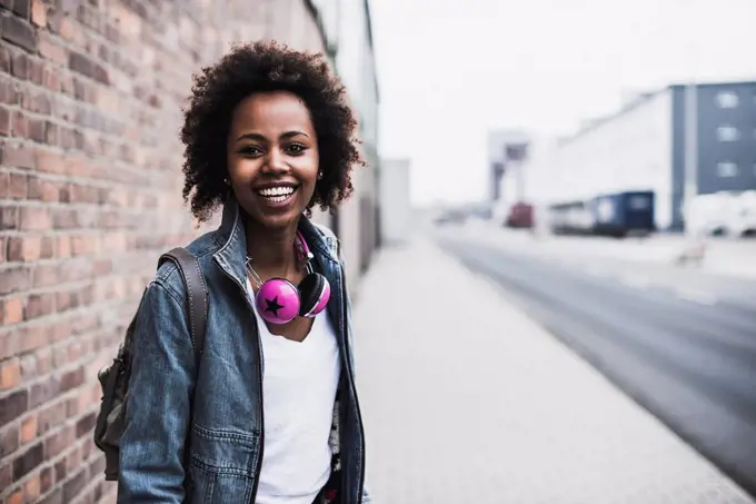 Portrait of smiling young woman with headphones and backpack standing on pavement