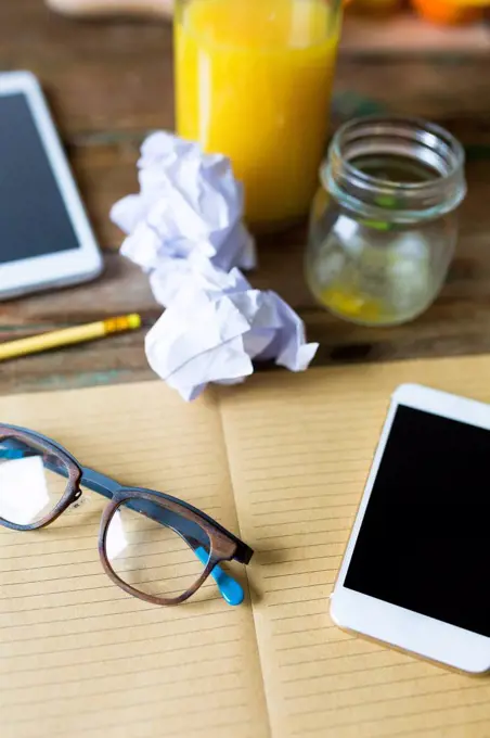 Glass bottle of orange juice, empty glass, notepad, spectacles, tablet, cell phone and crumpled paper