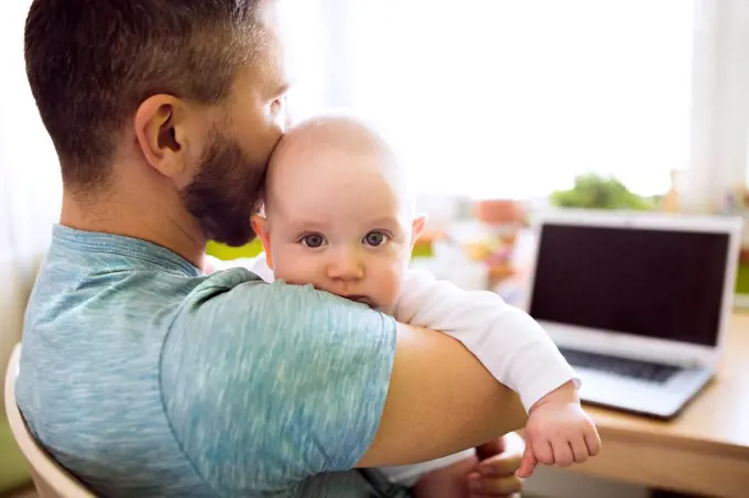 Father holding baby son at home with laptop on table