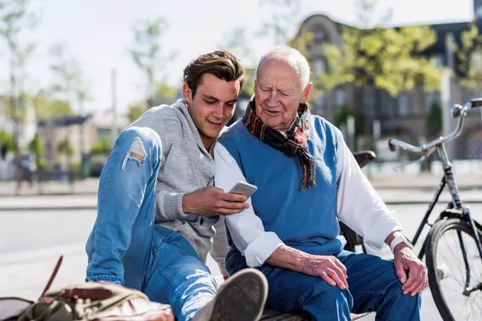 Senior man and adult grandson on a bench looking at cell phone