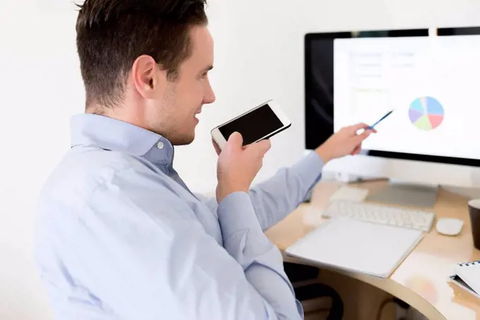 Businessman with smartphone analysing pie chart on computer screen