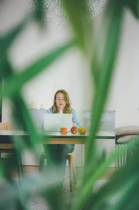 Young woman with closed eyes sitting at table with apples and laptop