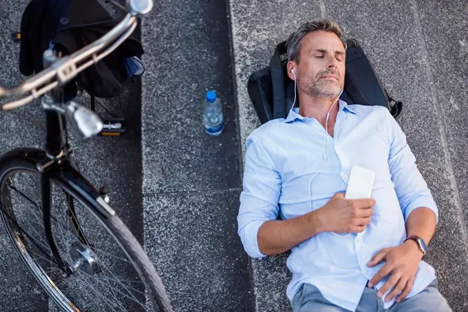 Man with closed eyes lying on stairs with cell phone and earbuds next to bicycle