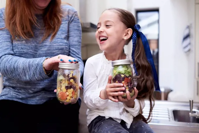 Laughing little girl with preserving jar of mixed salad sitting beside her mother