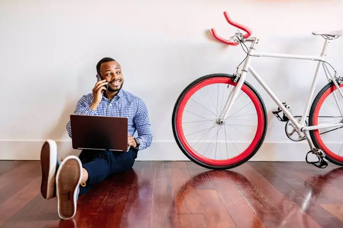 Smiling man on cell phone sitting on wooden floor with bicycle next to him