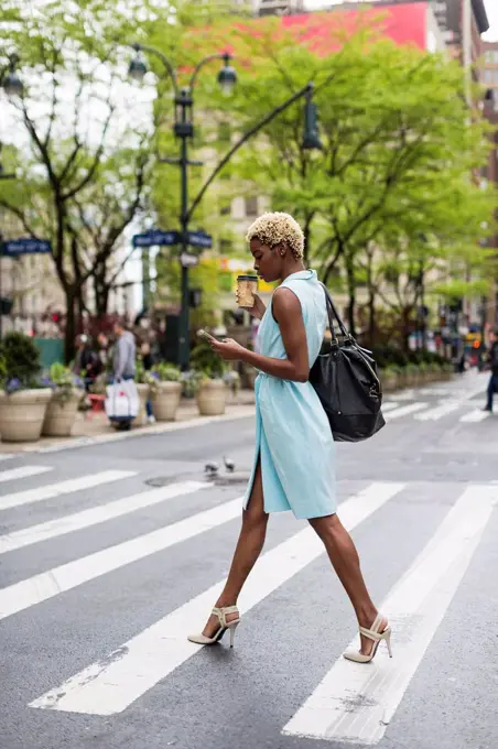USA, New York, young blonde african-american woman with cup of coffee and smart phone crossing street