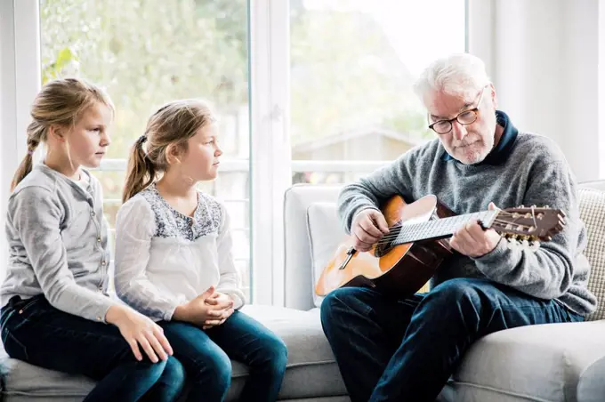 Two girls sitting on sofa listening to grandfather playing guitar