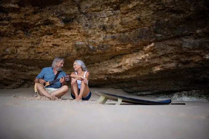 Handsome senior couple sitting on the beach, man playing guitar