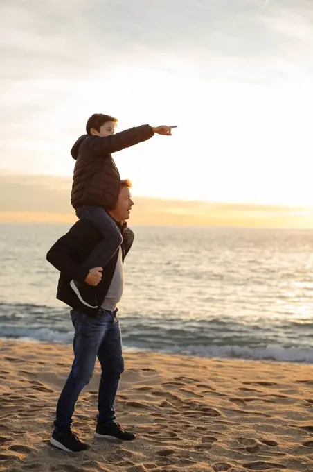 Father carrying son piggyback on the beach at sunset pointing finger