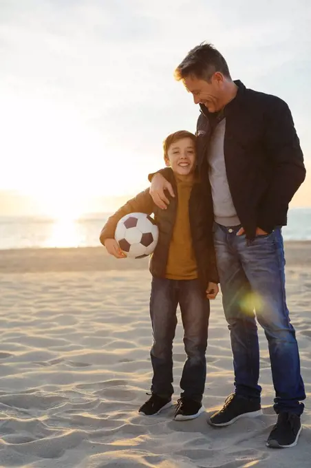 Father embracing son with football on the beach at sunset