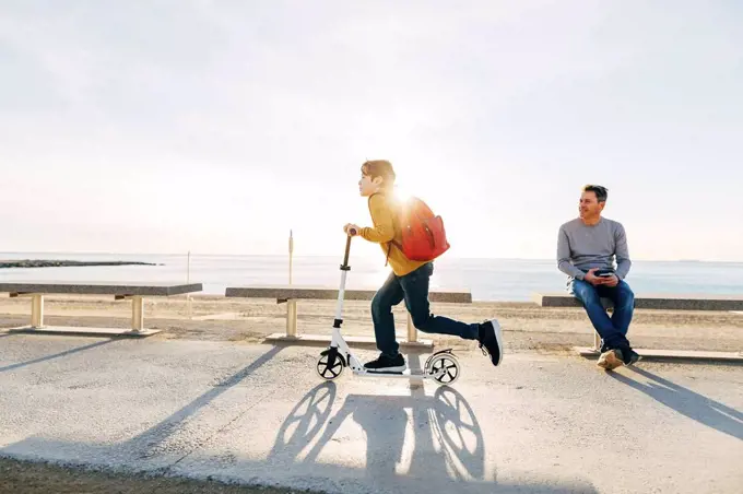 Father watching son riding scooter on beach promenade at sunset