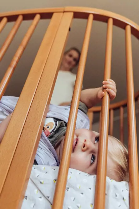 Baby lying in crib with open eyes, mother watching in background