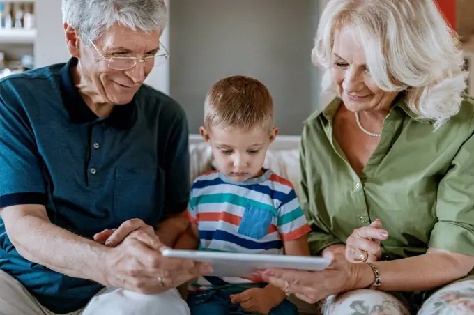 Grandparents and grandson at home sitting on couch sharing tablet