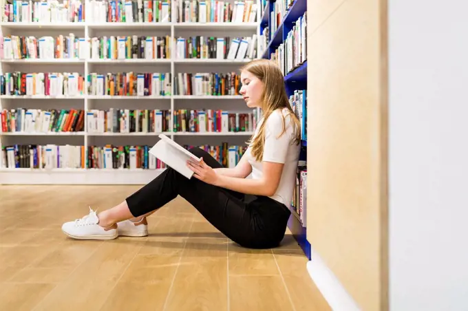 Teenage girl sitting on the floor in a public library reading book