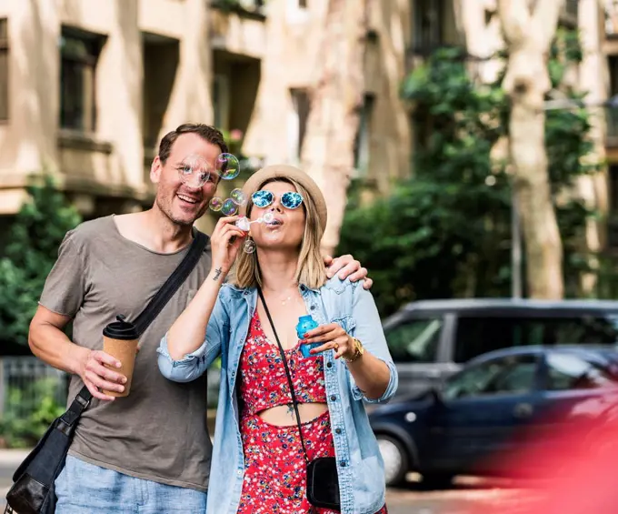 Happy couple with coffee to go blowing soap bubbles in the city