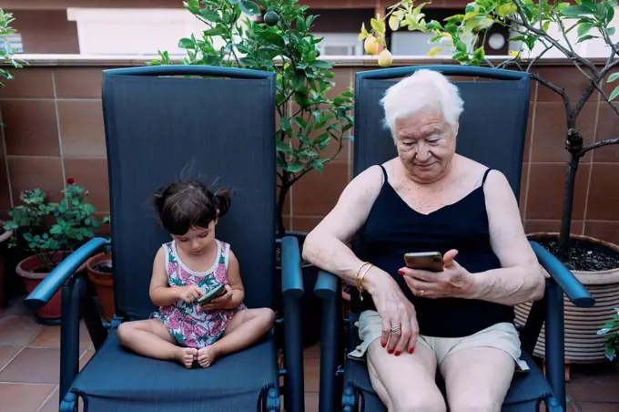 Grandmother and baby girl sitting side by side on the terrace using mobile phones