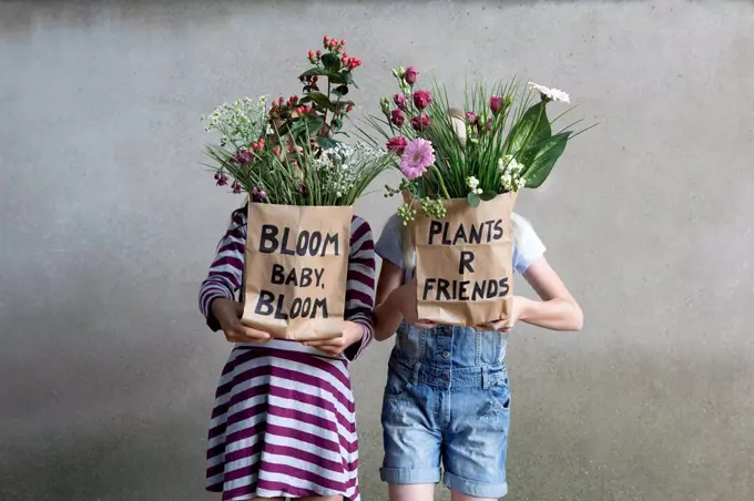 Two girls standing side by side hiding behind paper bags with flowers