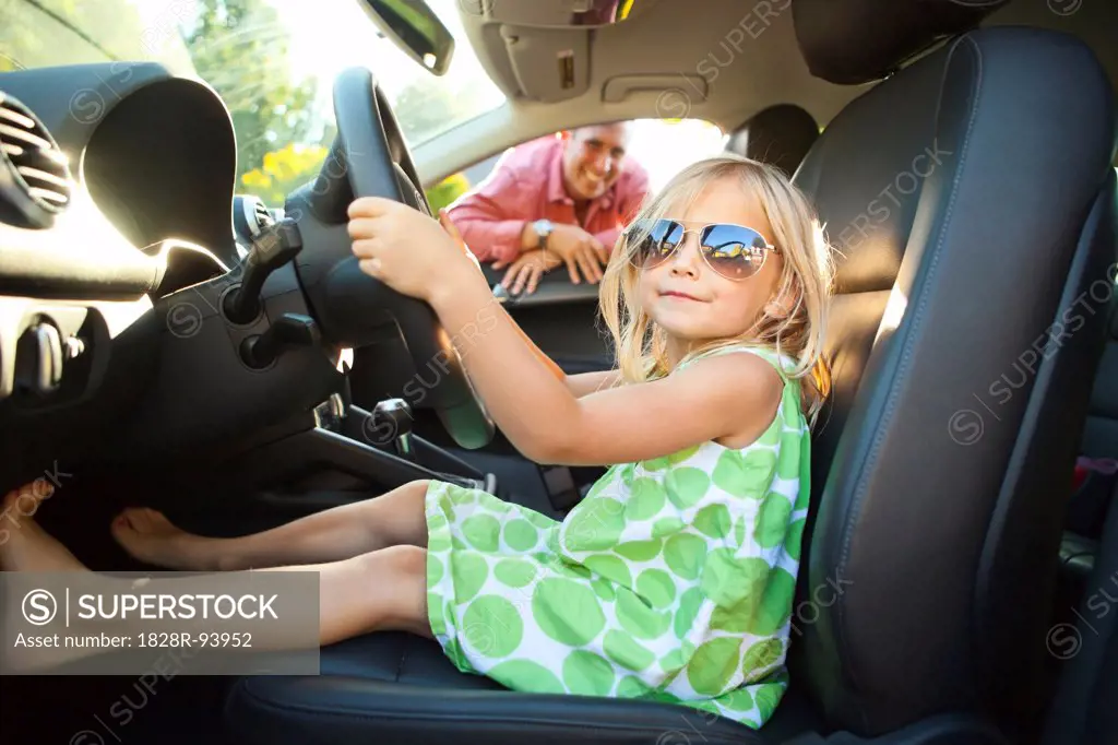 Portrait of little girl sitting in driver's seat of car, pretending to be old enough to drive as her smiling father watches on on a sunny summer evening in Portland, Oregon, USA