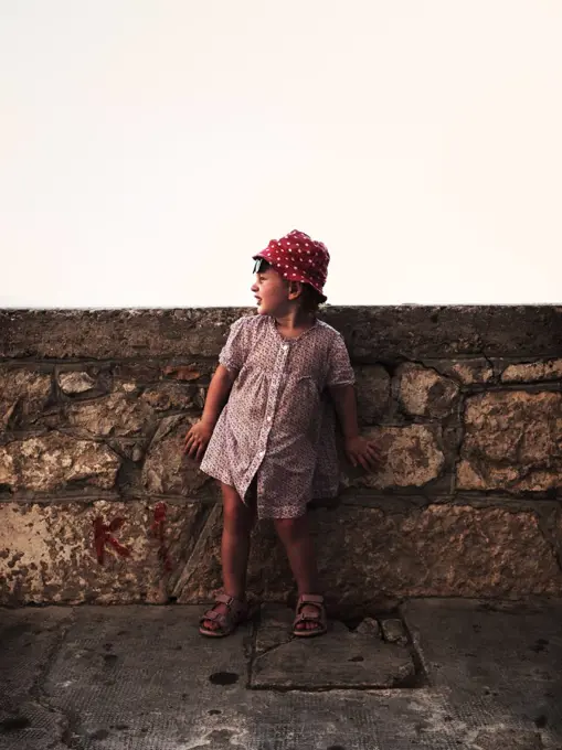 Young Girl in Red Hat and Print Dress Leaning Against Stone Wall, Croatia