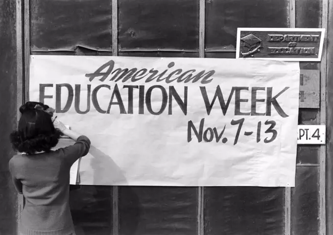 Woman hanging Sign promoting American Education Week to Wall of Education Department office, Manzanar Relocation Center, California, USA, Ansel Adams, Manzanar War Relocation Center Collection, 1943 