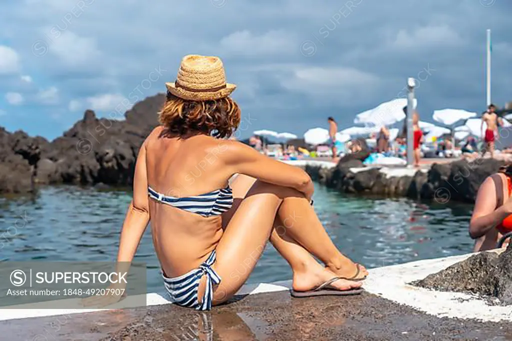 Porto Moniz seaside village, young tourist in hat and bikini sitting in the natural pools, Madeira