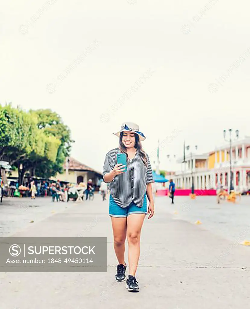 Smiling young tourist in hat walking with cell phone on the street. Female tourist texting phone in the square of Granada