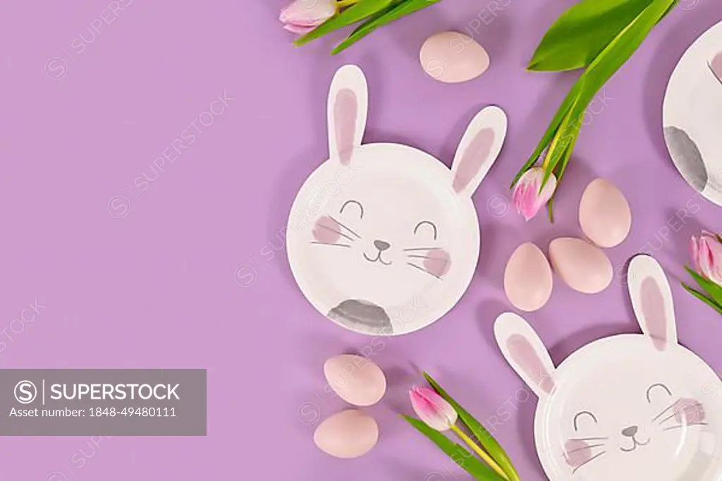 Easter party flat lay with bunny shaped paper plates, eggs and tulip flowers on violet background with copy space