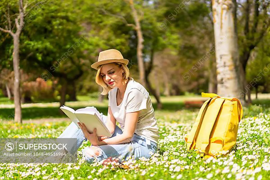 A young blonde girl in a hat reading a book in spring in a park in the city, vacations next to nature and next to daisies, sitting on the grass