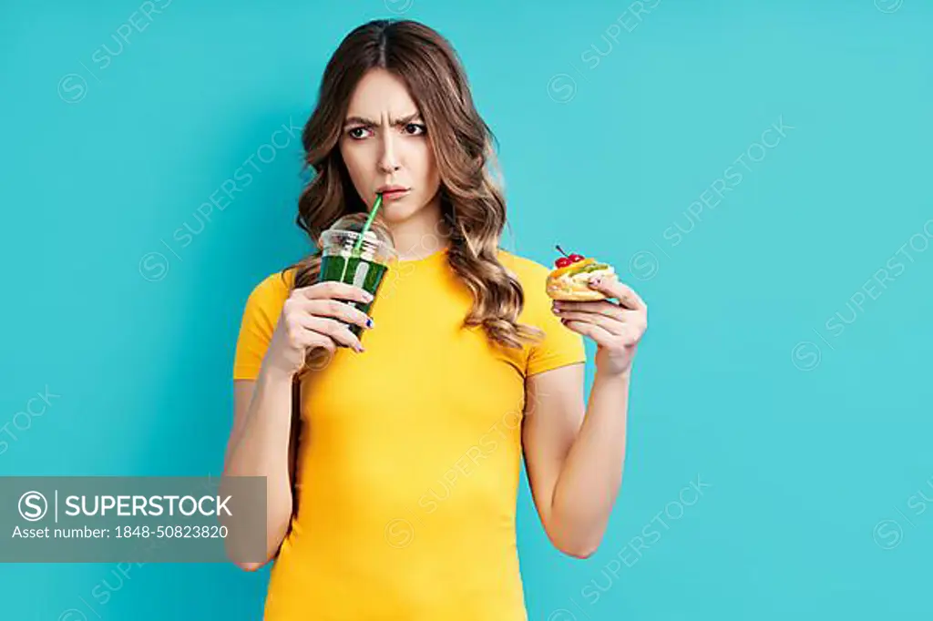 Unhappy woman on dieting drinking detox juice holding cake in hand choosing healthy food. Diet concept. eating lifestyle