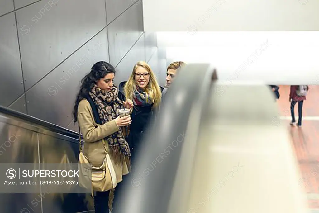 Three young adult women dressed in coat and scarves moving up on escalator in building with copy space on side