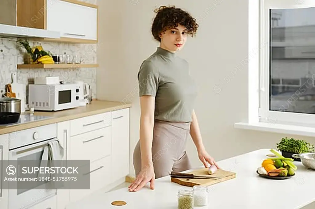 Woman stands in a well-lit modern kitchen preparing ingredients on a table. Young woman is ready to cook healthy food