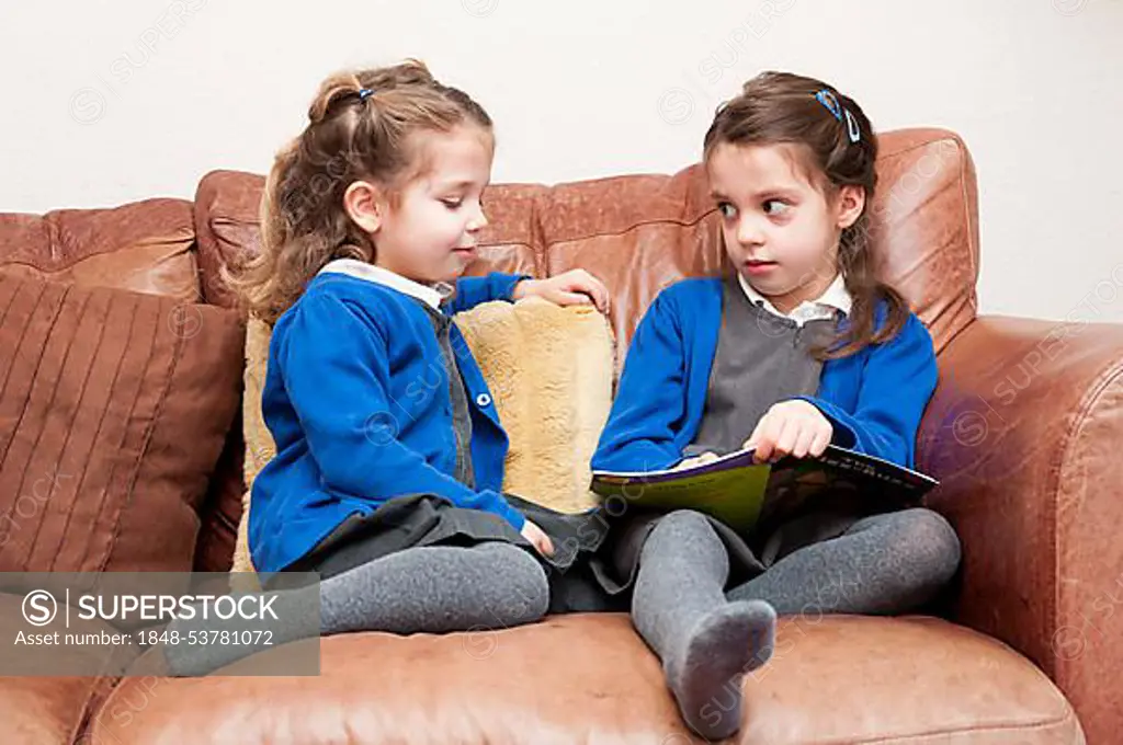 Young sisters in Primary School uniform reading a book together, sitting on a sofa, United Kingdom, Europe