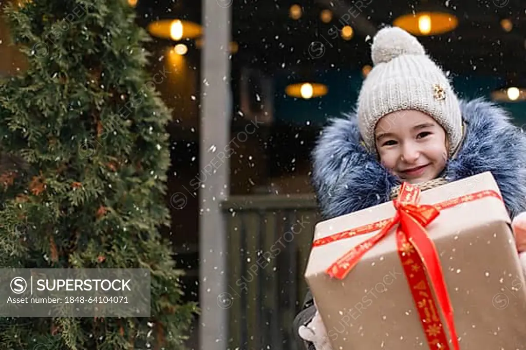 Portrait of joyful girl with a gift box for Christmas on a city street in winter with snow on a festive market with decorations and lights. Warm clothes, knitted hat, scarf and fur. Copy space