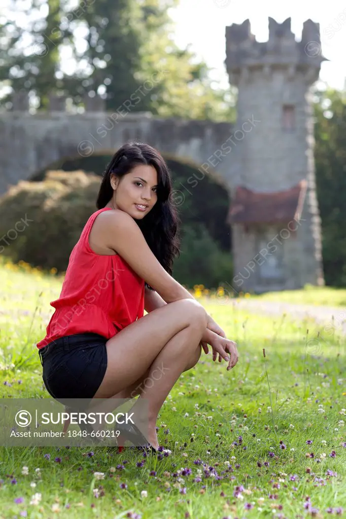 Young woman in a red top, black hot pants and high heels posing on a meadow in front of a castle tower