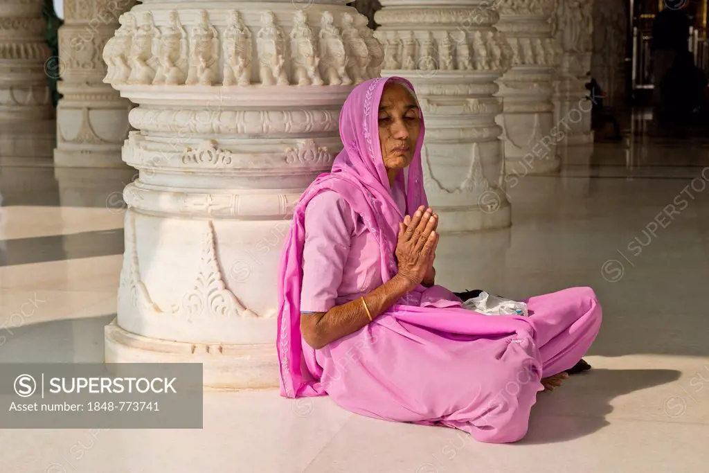 Old Indian woman in a sari sitting on the floor in prayer, in front of an ornate marble column, at Shri Swaminarayan Mandir, a Hindu temple of the Swa...