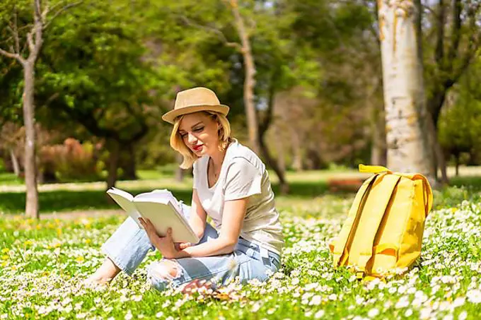 A young blonde girl in a hat reading a book in spring in a park in the city, vacations next to nature and next to daisies, sitting on the grass