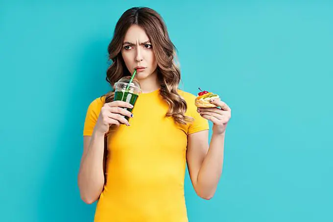 Unhappy woman on dieting drinking detox juice holding cake in hand choosing healthy food. Diet concept. eating lifestyle