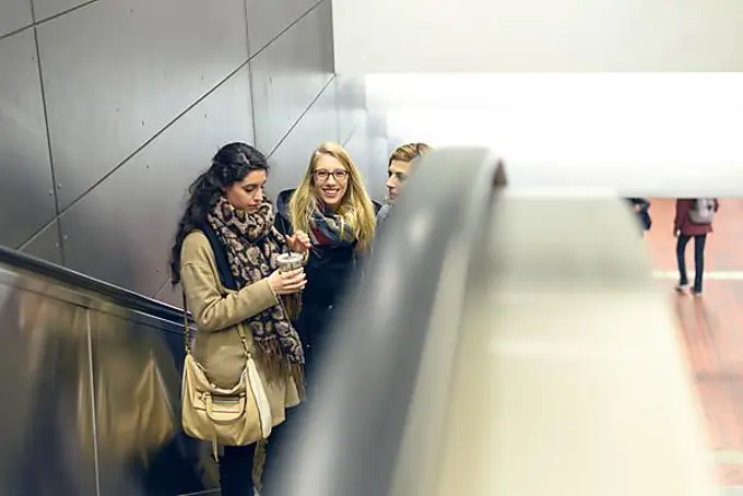 Three young adult women dressed in coat and scarves moving up on escalator in building with copy space on side