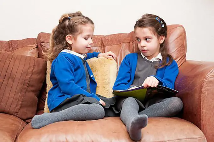 Young sisters in Primary School uniform reading a book together, sitting on a sofa, United Kingdom, Europe