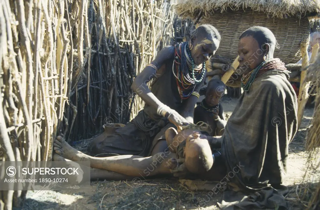 Uganda, Karamoja, Female Witch Doctor Or Amuron Using Sysal To Suck Out Evil From Child Affected By The Evil Eye.  Karamojong Women Paint Their Faces With Clay To Prevent Disease.