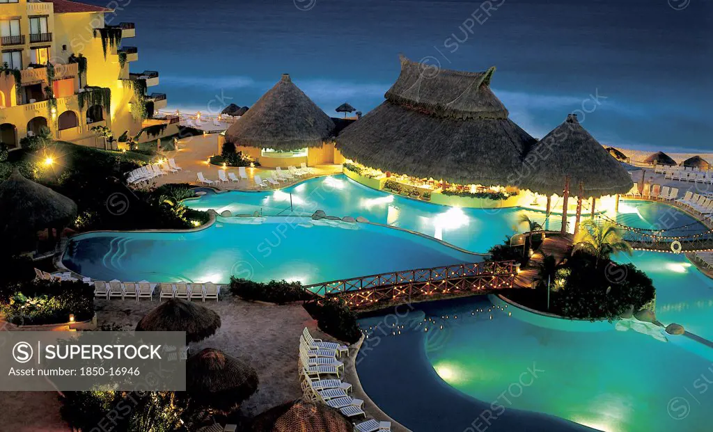 Mexico, Jalisco ,  Puerto Vallarta, 'A Swimming Pool Surrounded By Sun Loungers, Wooden Thatched Huts And Bridges Lit Up At Night. Hotel Right Next To Beach.'