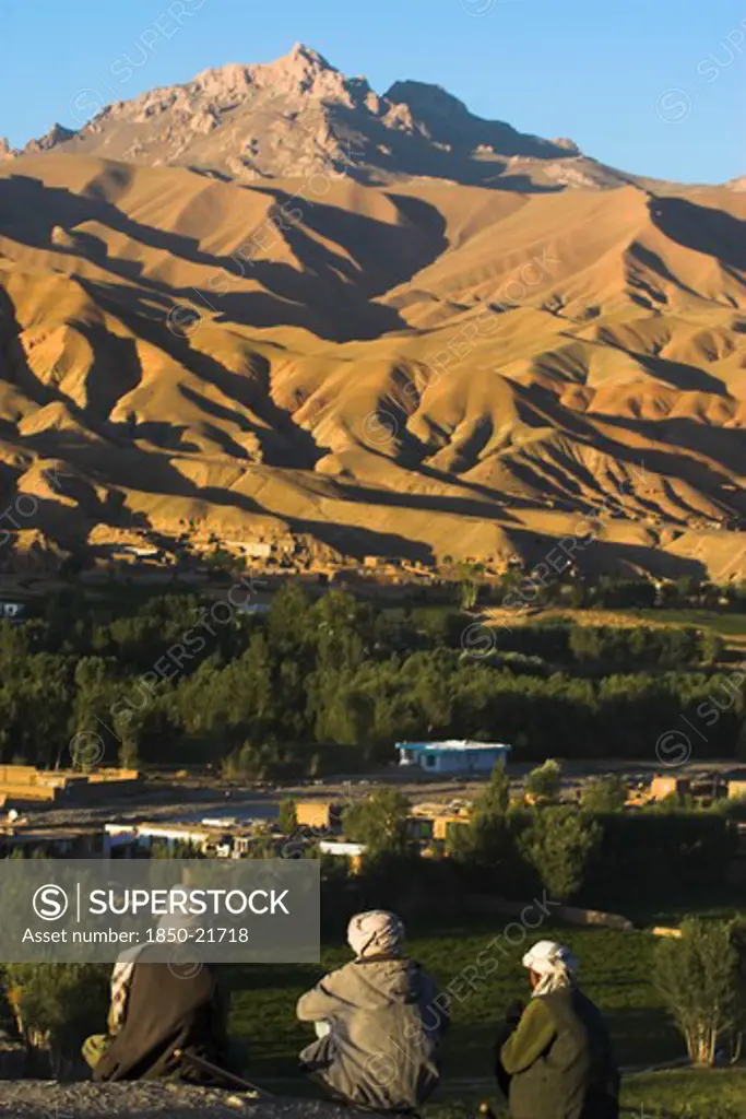 Afghanistan, Bamiyan Province, Bamiyan, Men Sit Above Bamiyan Village As The Late Afternoon Sun Glows On Mountains Near The Empty Niche Where The Famous Carved Budda Once Stood (Destroyed By The Taliban In 2001)